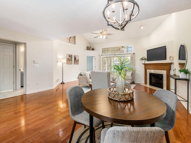 dining room featuring baseboards, light wood finished floors, lofted ceiling, a fireplace with flush hearth, and ceiling fan with notable chandelier