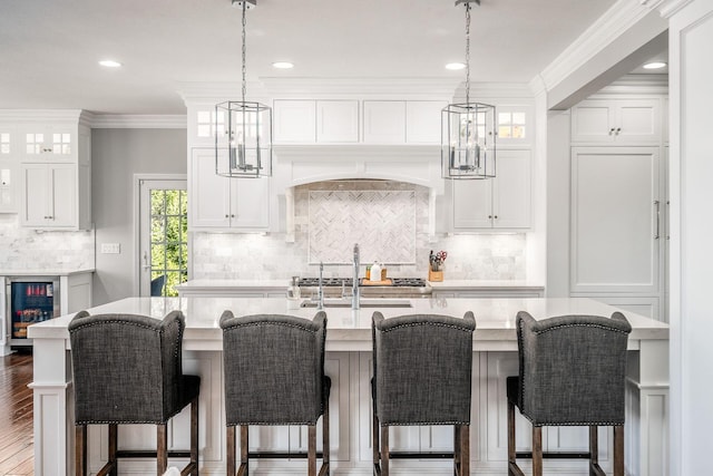 kitchen featuring beverage cooler, an island with sink, ornamental molding, white cabinets, and glass insert cabinets