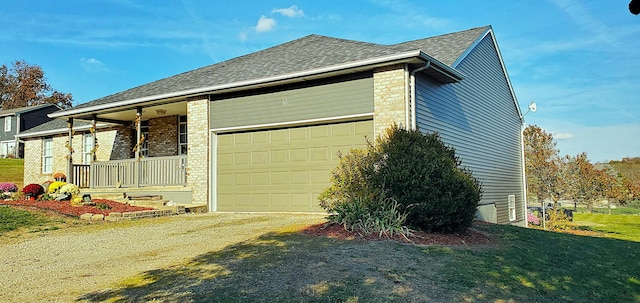 view of side of home featuring a porch, dirt driveway, roof with shingles, an attached garage, and brick siding