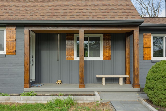 view of exterior entry with covered porch, brick siding, and roof with shingles