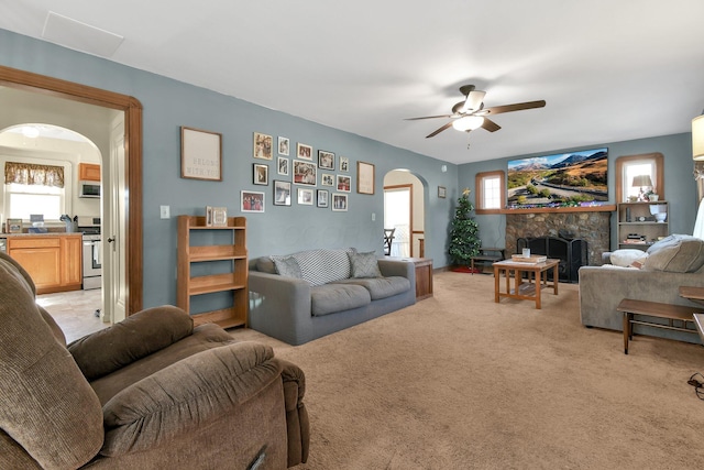 living room featuring a stone fireplace, ceiling fan, arched walkways, and light carpet