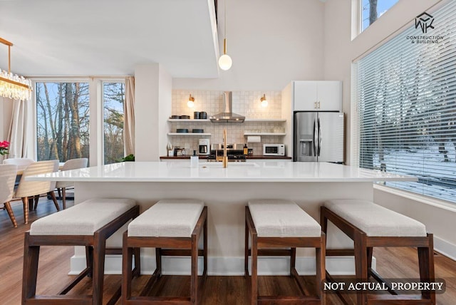 kitchen featuring white microwave, stainless steel fridge with ice dispenser, dark wood-style flooring, decorative backsplash, and wall chimney range hood