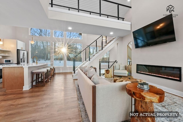 living room featuring light wood-style flooring, a glass covered fireplace, baseboards, a towering ceiling, and stairs