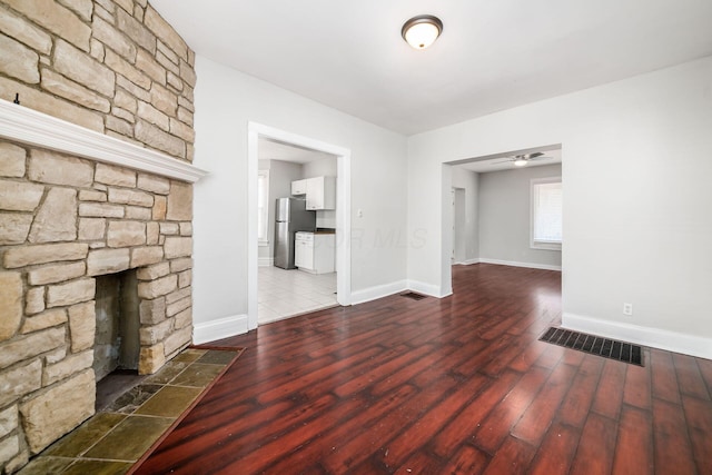 unfurnished living room with wood finished floors, visible vents, baseboards, ceiling fan, and a stone fireplace