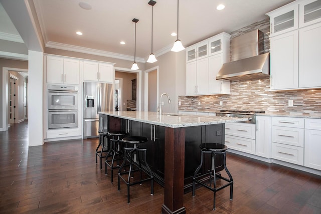 kitchen featuring an island with sink, backsplash, appliances with stainless steel finishes, a breakfast bar area, and wall chimney range hood