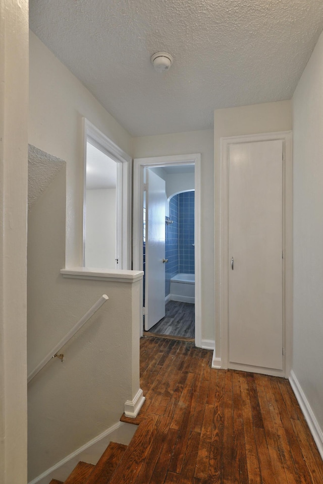 hallway with an upstairs landing, a textured ceiling, baseboards, and dark wood-style flooring