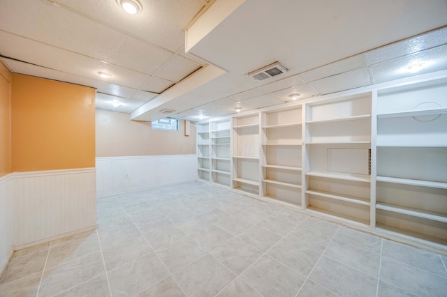 below grade area featuring tile patterned floors, visible vents, built in shelves, a drop ceiling, and wainscoting