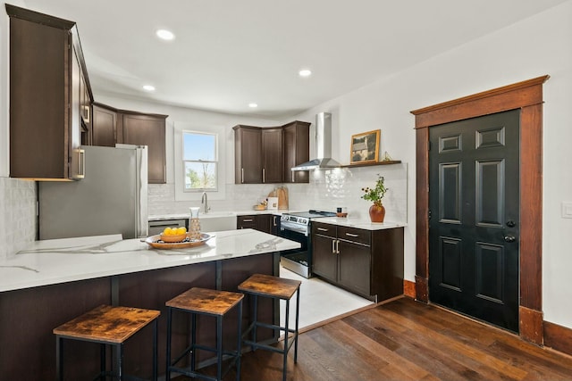 kitchen featuring wood finished floors, a peninsula, stainless steel appliances, dark brown cabinetry, and wall chimney range hood