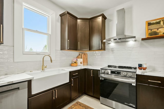 kitchen with a sink, light stone counters, dark brown cabinetry, appliances with stainless steel finishes, and wall chimney range hood