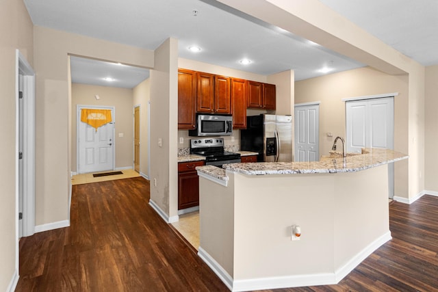 kitchen with light stone counters, baseboards, a center island with sink, dark wood finished floors, and stainless steel appliances