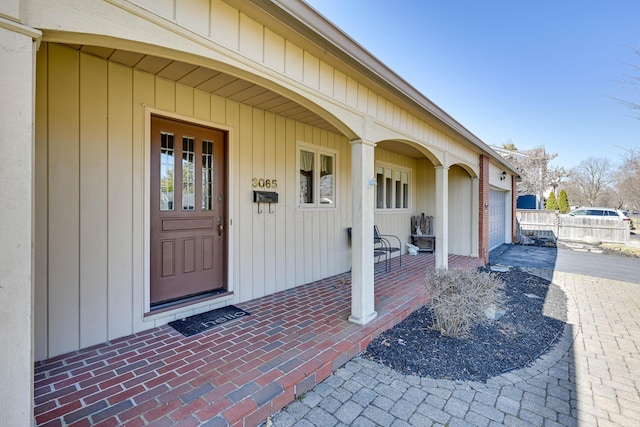 doorway to property with brick siding, covered porch, an attached garage, and fence