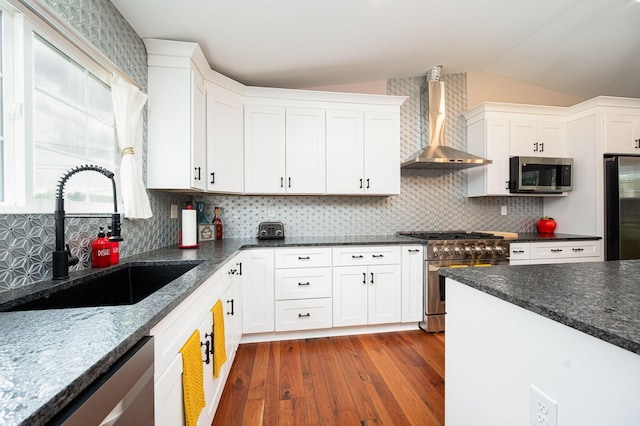 kitchen with a sink, stainless steel appliances, vaulted ceiling, white cabinetry, and wall chimney exhaust hood