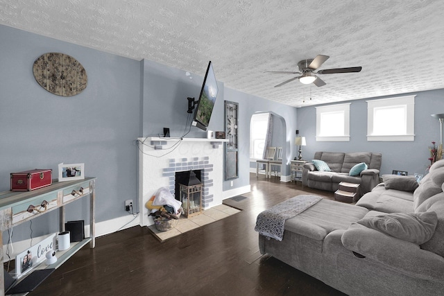 living room featuring a brick fireplace, ceiling fan, wood finished floors, arched walkways, and a textured ceiling