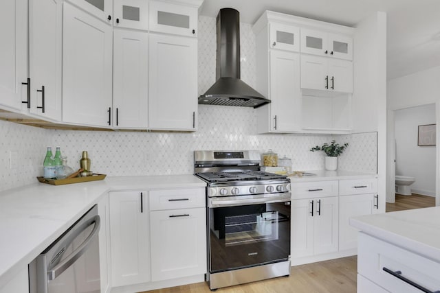 kitchen with stainless steel appliances, white cabinetry, wall chimney range hood, light wood-type flooring, and backsplash