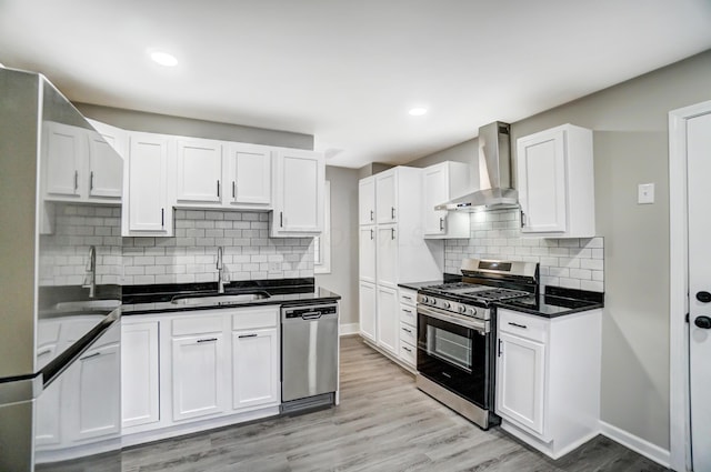 kitchen with wall chimney range hood, light wood-style flooring, appliances with stainless steel finishes, white cabinets, and a sink