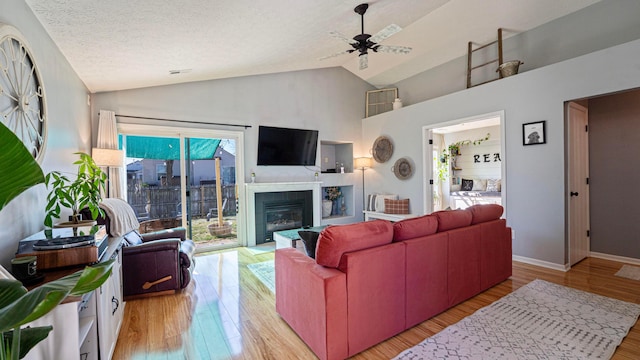 living area featuring high vaulted ceiling, light wood-style flooring, ceiling fan, a textured ceiling, and a glass covered fireplace