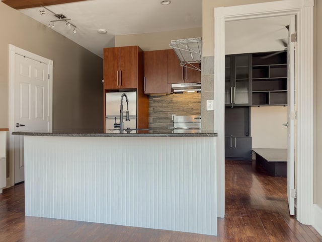 kitchen featuring a peninsula, a sink, decorative backsplash, under cabinet range hood, and brown cabinets