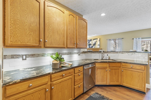 kitchen with light wood finished floors, dark stone countertops, a peninsula, stainless steel dishwasher, and a sink