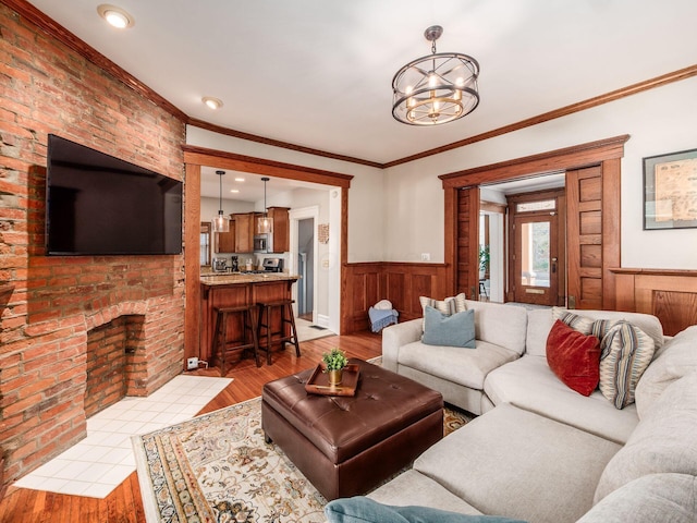 living room featuring ornamental molding, light wood-style floors, wainscoting, a brick fireplace, and a chandelier