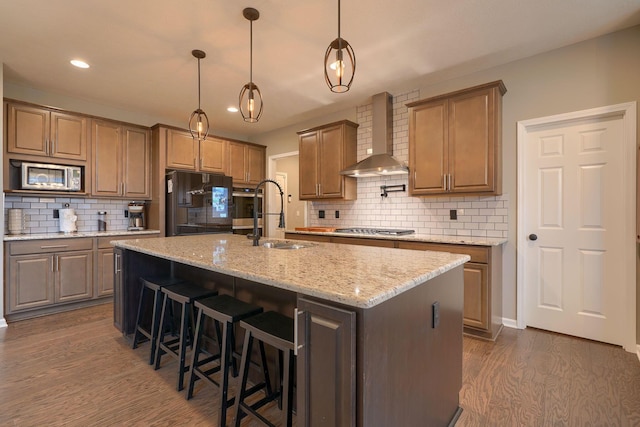 kitchen featuring a center island with sink, dark wood-style floors, stainless steel appliances, wall chimney exhaust hood, and a sink