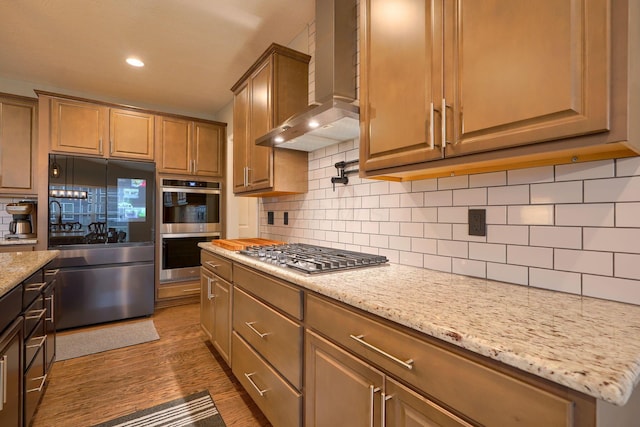 kitchen with dark wood-type flooring, backsplash, stainless steel appliances, wall chimney exhaust hood, and light stone countertops