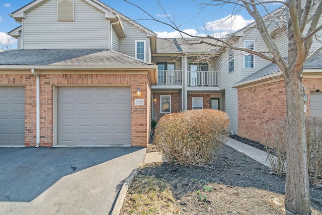 multi unit property featuring a balcony, roof with shingles, an attached garage, aphalt driveway, and brick siding