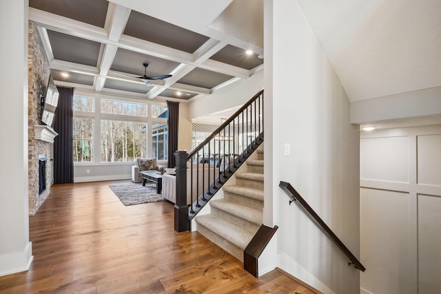 stairway with wood finished floors, a ceiling fan, coffered ceiling, beam ceiling, and a stone fireplace