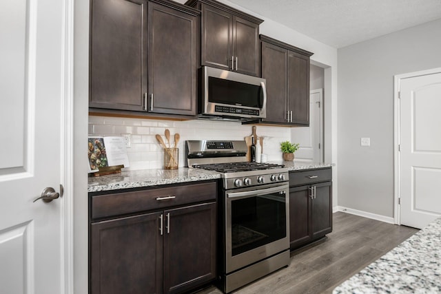 kitchen with backsplash, dark wood finished floors, dark brown cabinetry, appliances with stainless steel finishes, and light stone countertops