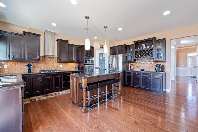 kitchen with a sink, dark stone counters, appliances with stainless steel finishes, and wall chimney range hood