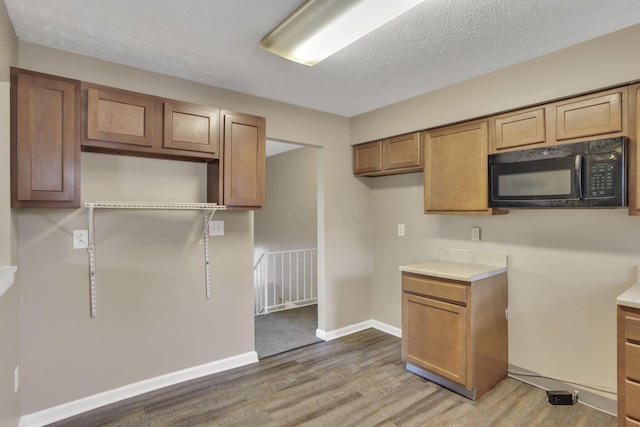 kitchen with black microwave, wood finished floors, brown cabinetry, baseboards, and a textured ceiling