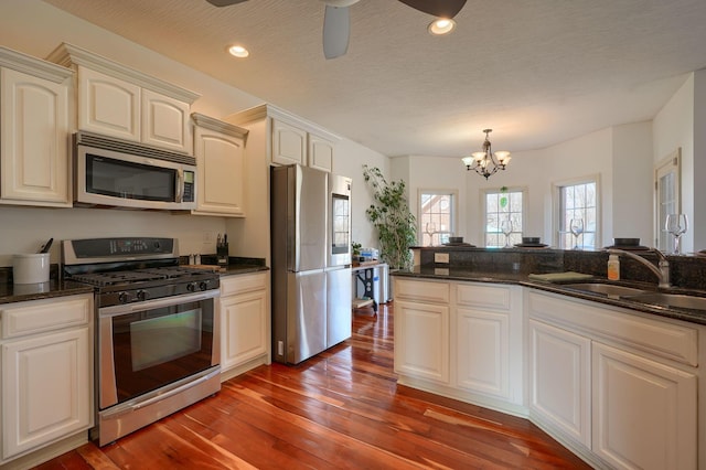 kitchen with ceiling fan with notable chandelier, a sink, hardwood / wood-style floors, stainless steel appliances, and dark stone counters