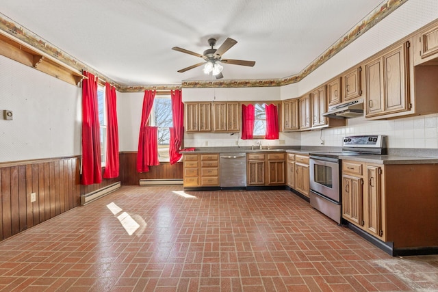 kitchen with a wainscoted wall, under cabinet range hood, appliances with stainless steel finishes, wood walls, and baseboard heating