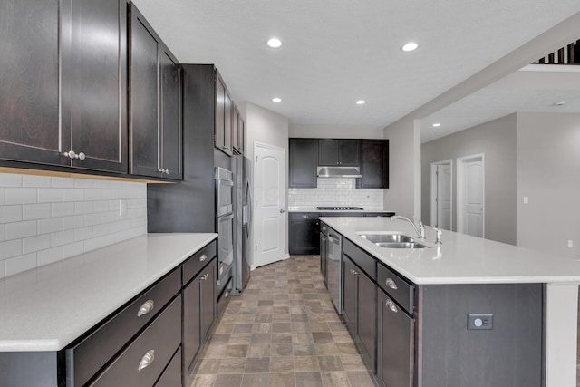 kitchen with light countertops, a sink, appliances with stainless steel finishes, a center island with sink, and under cabinet range hood
