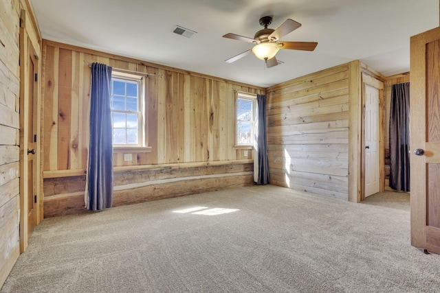 carpeted empty room featuring wooden walls, a healthy amount of sunlight, visible vents, and ceiling fan