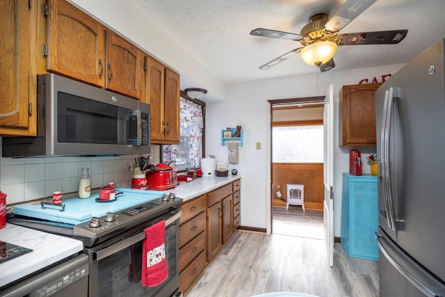 kitchen featuring backsplash, appliances with stainless steel finishes, brown cabinetry, light wood finished floors, and ceiling fan