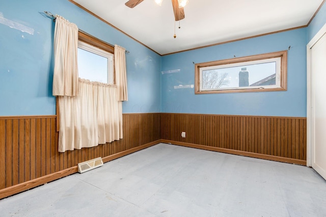 unfurnished room featuring wooden walls, visible vents, ceiling fan, ornamental molding, and wainscoting