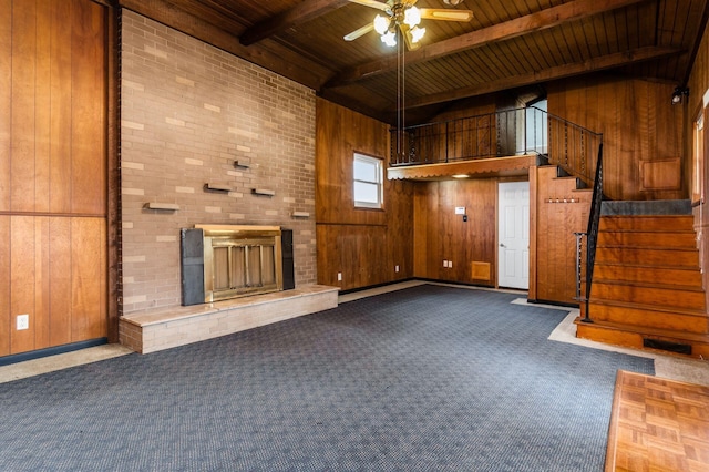 unfurnished living room with a brick fireplace, beamed ceiling, wood walls, carpet floors, and a ceiling fan