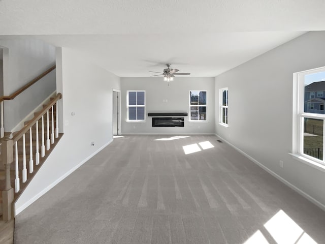 unfurnished living room featuring baseboards, carpet, stairs, a glass covered fireplace, and a ceiling fan