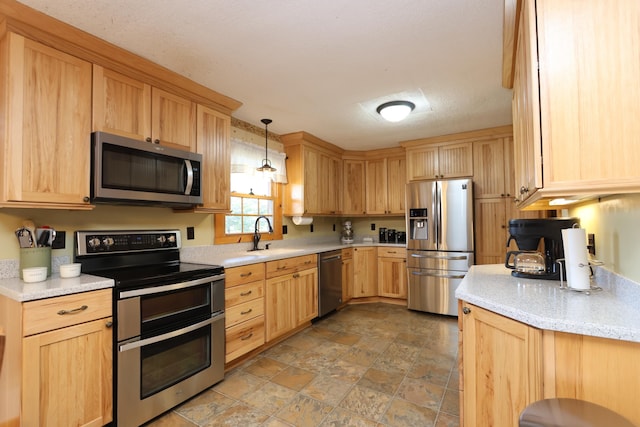 kitchen with light brown cabinets, a sink, stone finish floor, appliances with stainless steel finishes, and decorative light fixtures