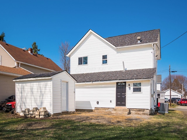 back of house with an outbuilding, a lawn, central AC, and roof with shingles