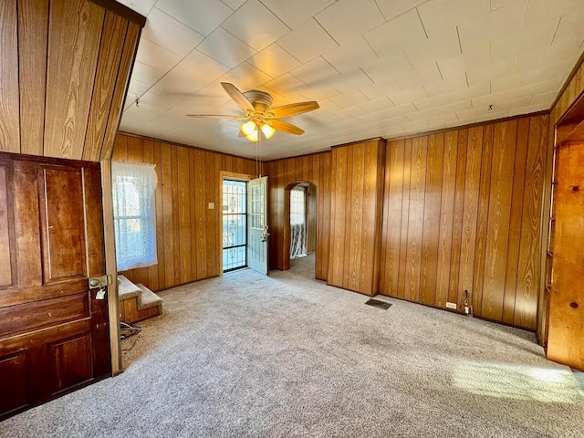 carpeted empty room featuring visible vents, arched walkways, wood walls, and a ceiling fan