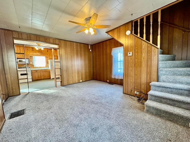 unfurnished living room with visible vents, a ceiling fan, wooden walls, carpet, and stairs