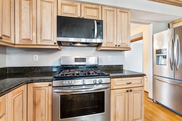 kitchen with light brown cabinetry, light wood-style flooring, stainless steel appliances, and dark stone countertops