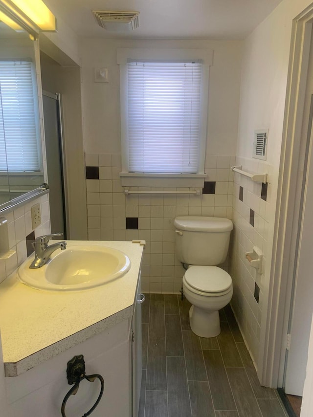 full bath with a shower with door, visible vents, toilet, vanity, and tile walls