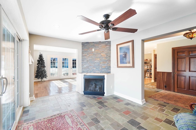 living area featuring baseboards, ceiling fan, a stone fireplace, stone tile flooring, and french doors