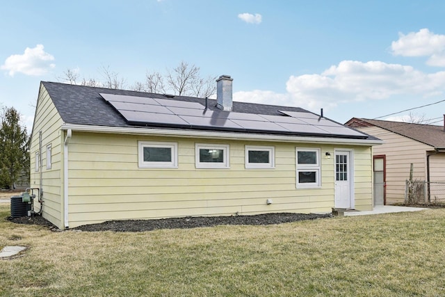 rear view of property with solar panels, a shingled roof, central air condition unit, a lawn, and a chimney