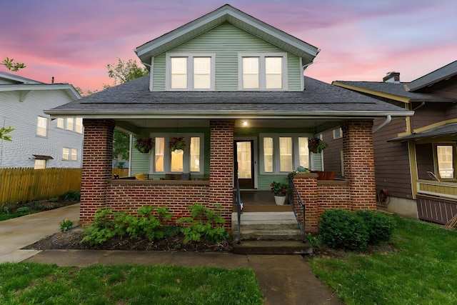 bungalow-style house featuring roof with shingles, brick siding, covered porch, and fence