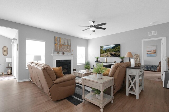 living room with a ceiling fan, visible vents, light wood finished floors, a textured ceiling, and a glass covered fireplace