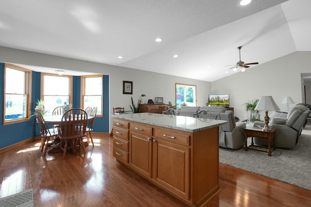 kitchen with ceiling fan, light stone counters, dark wood-style floors, vaulted ceiling, and a center island