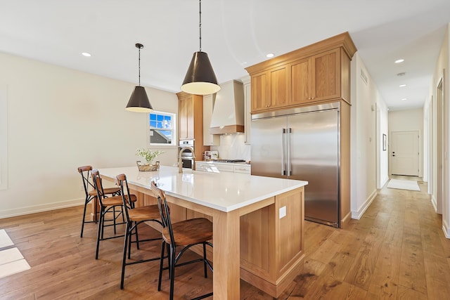 kitchen featuring a center island with sink, light countertops, light wood-style flooring, appliances with stainless steel finishes, and wall chimney exhaust hood